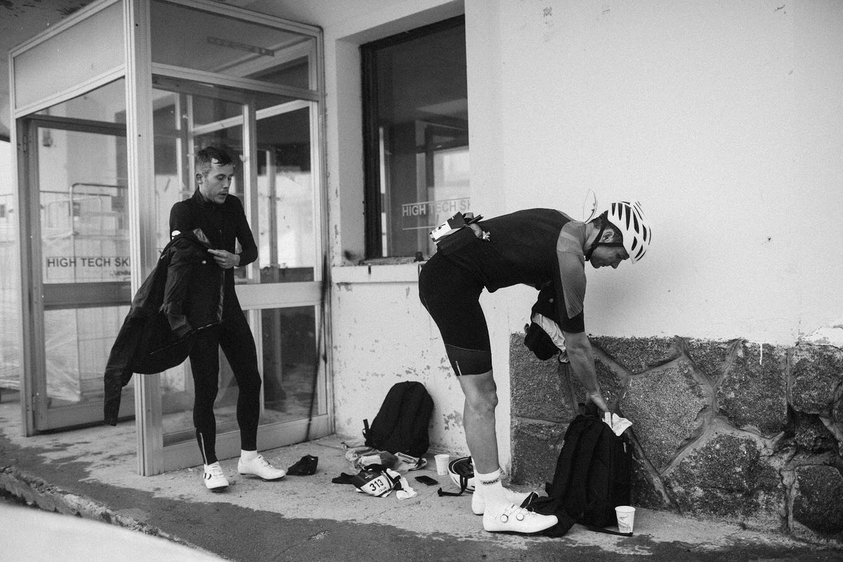 Black and white street photograph of two cyclists preparing outside a building. One cyclist in a black cycling kit stands near the glass entrance holding a jacket, while another cyclist in a black jersey and shorts bends over to pack gear on the ground beside a stone base.